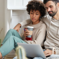 Photo of mixed race partners enjoy day off and spare time, embrace and watch tutorial video together on laptop computer. Handsome guy in casual sweatshirt hugs wife, pose on floor in apartment