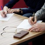 An adult woman and a man, sitting at a table in the office, fill out documents or forms. The concept of a marriage contract, agreement or divorce proceedings. Without a face.
