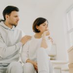 Photo of sad woman complains about problems, man calms, embraces wife, tired after bringing many boxes with things to their new home, wear casual clothes, being stressful. Family problems concept