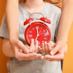 Hands of family with alarm clock on color background