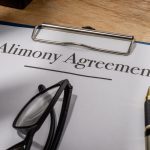 Clipboard with "Alimony Agreement" document, eyeglasses, and a pen on a wooden desk. The scene conveys a formal, legal atmosphere.