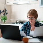 Senior woman working from home kitchen with laptop, documents, finances. Personal budgeting, financial literacy, pension plan, retirement lifestyle. Stress, money anxiety. Ageism in looking for job.