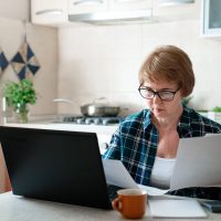 Senior woman working from home kitchen with laptop, documents, finances. Personal budgeting, financial literacy, pension plan, retirement lifestyle. Stress, money anxiety. Ageism in looking for job.