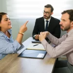 A woman in a striped shirt gestures while speaking passionately to two men in business attire across a table, conveying a serious discussion.