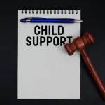 Notepad with "Child Support" text, blue pen on top, wooden judge’s gavel beside it, set against a black background. Legal, authoritative tone.