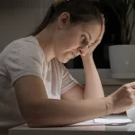 Woman sits at a table in a dimly lit room, reviewing documents with a focused, concerned expression. A plant and window are visible in the background.