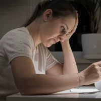 Woman sits at a table in a dimly lit room, reviewing documents with a focused, concerned expression. A plant and window are visible in the background.