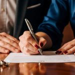 Couple hands signing a legal document on a table with wedding rings, symbolizing divorce or legal agreement.